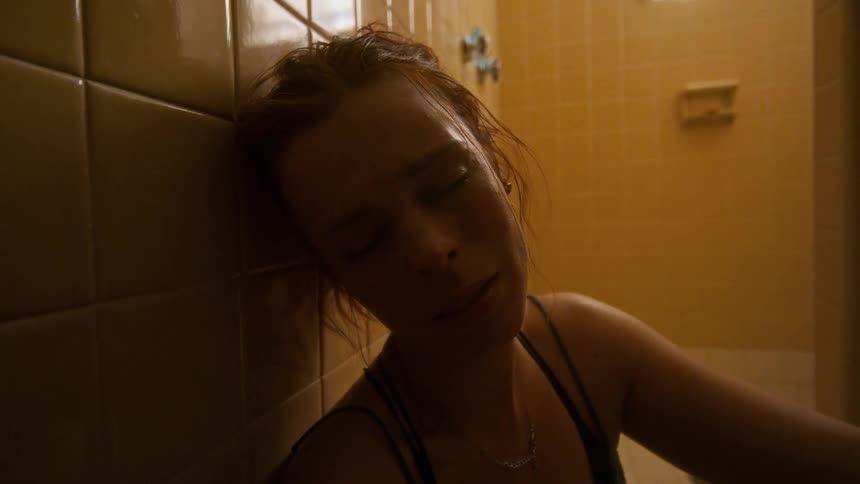 A distressed young woman with her eyes closed sits slumped against the wall in a dimly lit, tiled bathroom