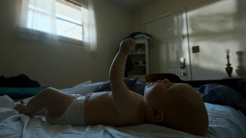 A mother gently picks up her baby from an unmade bed in a dimly lit and messy bedroom