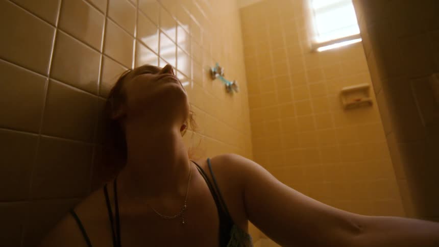 A troubled young woman sits passed out on the floor of a grimy shower in a dimly lit bathroom