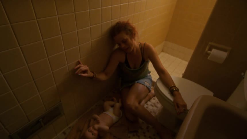 A distressed and exhausted mother slumps to the floor of a grimy bathroom next to her newborn baby