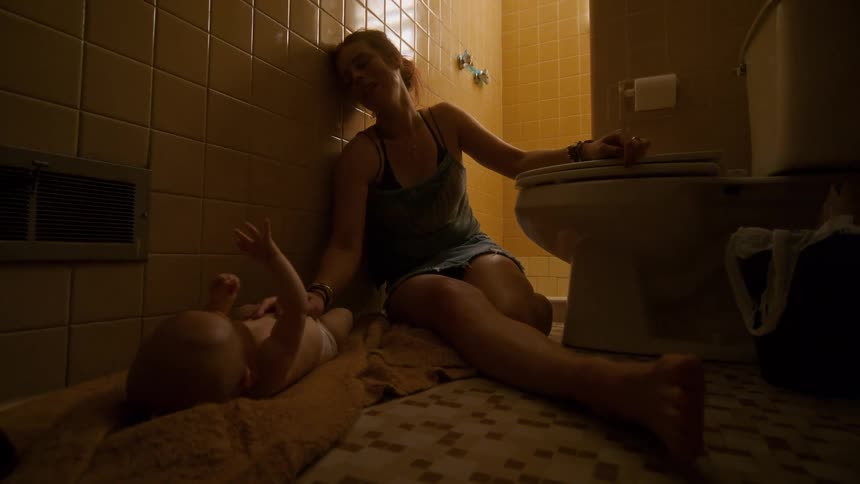 An exhausted and overwhelmed mother sits slumped against the wall on a dirty bathroom floor as her baby lays next to her on a towel