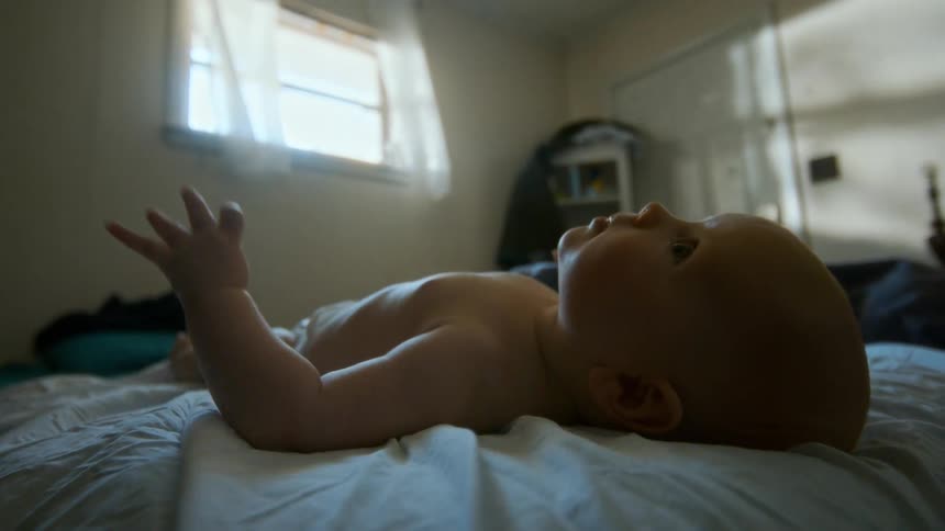 A peaceful baby boy wearing a diaper rests on a bed and moves his hands in a softly lit room
