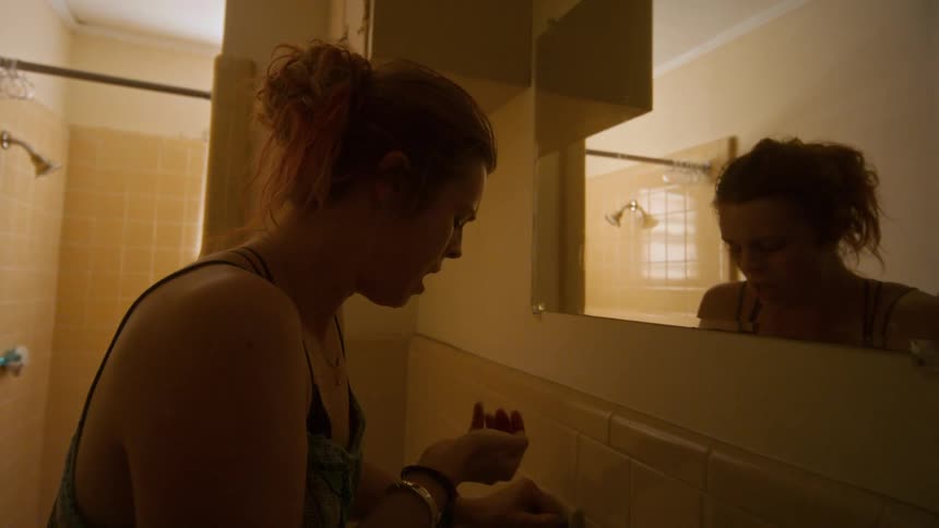A troubled woman takes a pill in a dimly lit bathroom and then stares at her somber reflection in the mirror