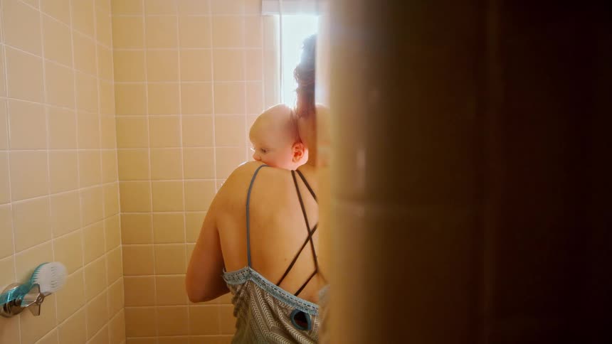 A distressed young mother holds her baby close for comfort while standing in a tiled shower stall
