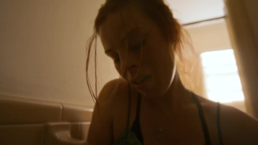 A distraught young woman with wet hair cries and slowly slumps down against the tiled wall of a dimly lit bathroom