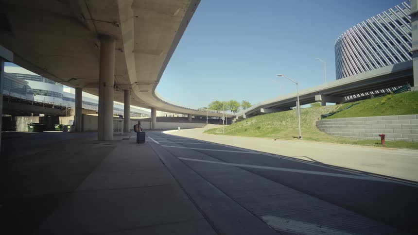 A lone traveler waits with his luggage under a vast concrete highway overpass at a modern transportation hub on a bright sunny day