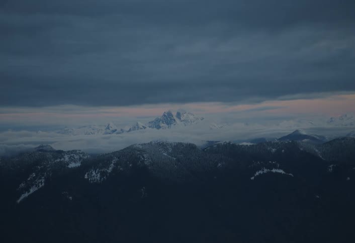 A moody and majestic aerial view slowly approaches snow-capped mountain peaks rising above a sea of clouds at twilight