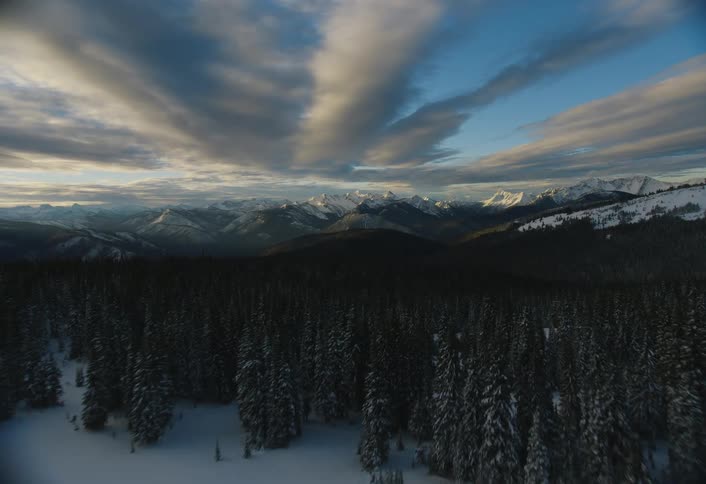 An epic aerial reveal flies over a snow-covered forest to showcase a majestic mountain range under a dramatic sky at sunrise