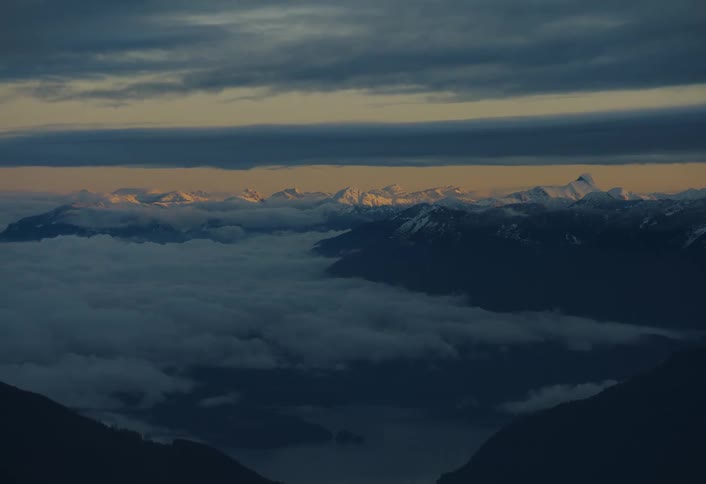 A stunning wide shot reveals a sea of clouds filling a valley with distant snow-capped mountains glowing at sunset