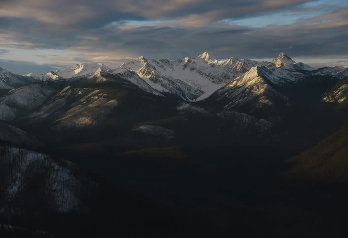 Golden sunrise light illuminates snowy mountain peaks above a vast, dark forested valley under a dramatic sky