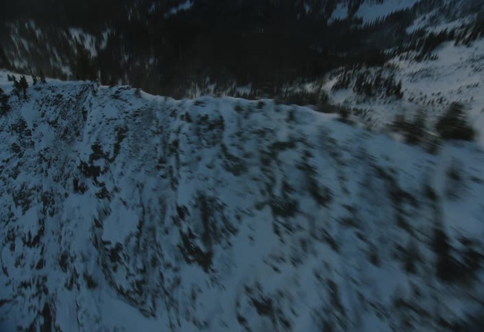 An aerial view crests a snowy mountain ridge at sunrise, then tilts down to fly over the vast, forested landscape below