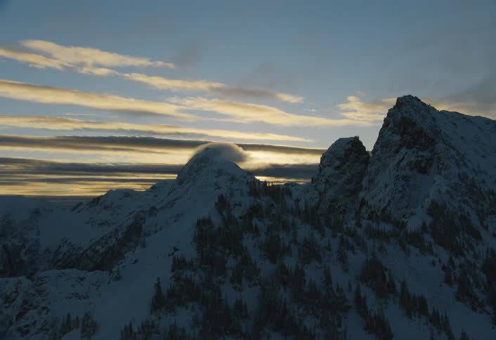 A dramatic aerial shot pulls back to reveal a vast, snow-covered mountain range silhouetted against a beautiful sunset sky