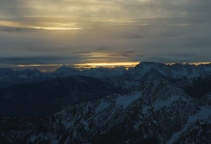 A sliver of golden sunset light breaks through dark, heavy clouds in a timelapse over a vast, snowy mountain range
