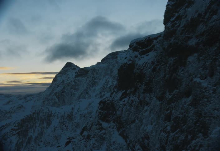A dramatic aerial shot flies alongside a rugged, snow-covered mountain face at dusk, revealing a vast alpine range