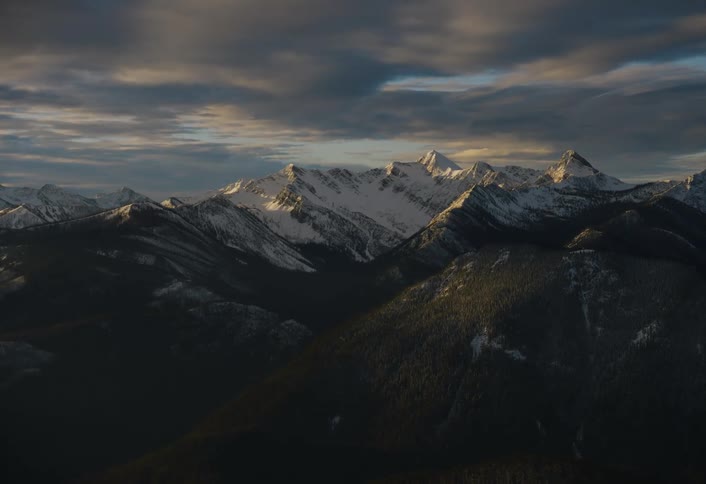A majestic wide shot of snow-capped mountains illuminated by the golden light of sunrise under a dramatic cloudy sky