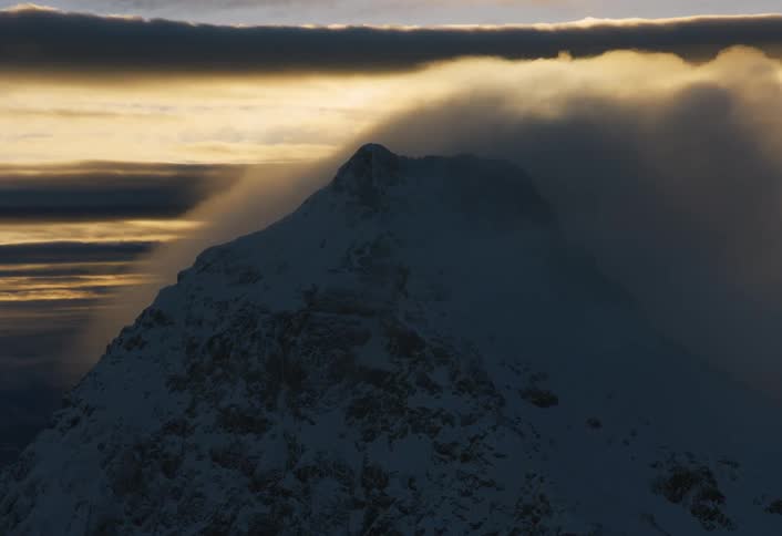 Fast-moving clouds envelop a snowy mountain peak during a dramatic sunset
