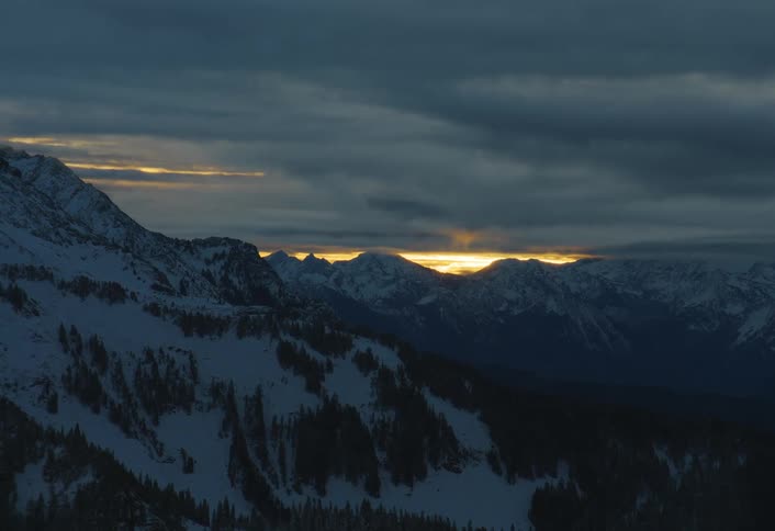 A slow aerial view reveals a vast, snow-covered mountain range as the sun sets behind the distant peaks under a dark, moody sky
