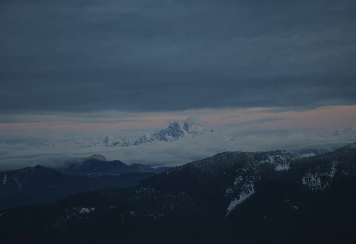 A serene aerial view of a dramatic, snow-capped mountain range rising above a sea of clouds at twilight