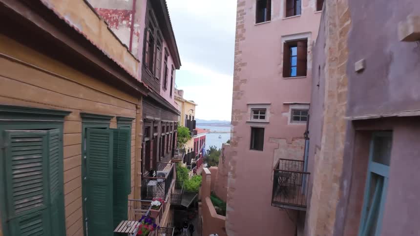 A slow aerial reveal shot flies through a narrow, historic alley in Chania, Crete, opening up to a beautiful view of the old Venetian harbor