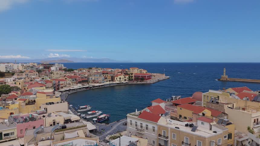 An aerial shot slowly moves towards the beautiful old venetian harbor of Chania, Crete, with its iconic lighthouse and colorful buildings