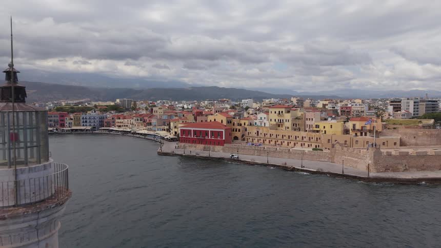 Aerial pullback shot revealing the historic lighthouse at the Old Venetian Harbour of Chania, Crete, under a cloudy sky