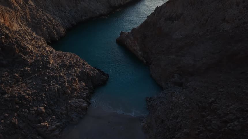 An aerial view flies over rocky cliffs to reveal a stunning hidden cove with turquoise water at sunset