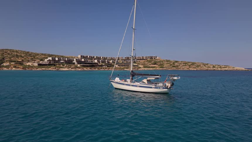 A sailboat is peacefully anchored in the stunningly clear turquoise waters of a coastal bay, with a modern resort visible on the shore