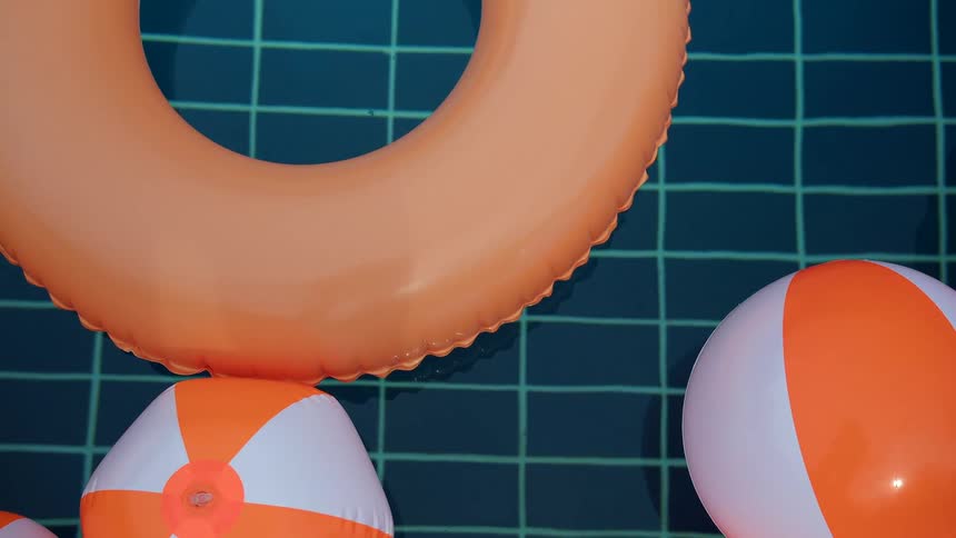 Overhead shot of orange and white pool floats and beach balls drifting in a tranquil tiled swimming pool