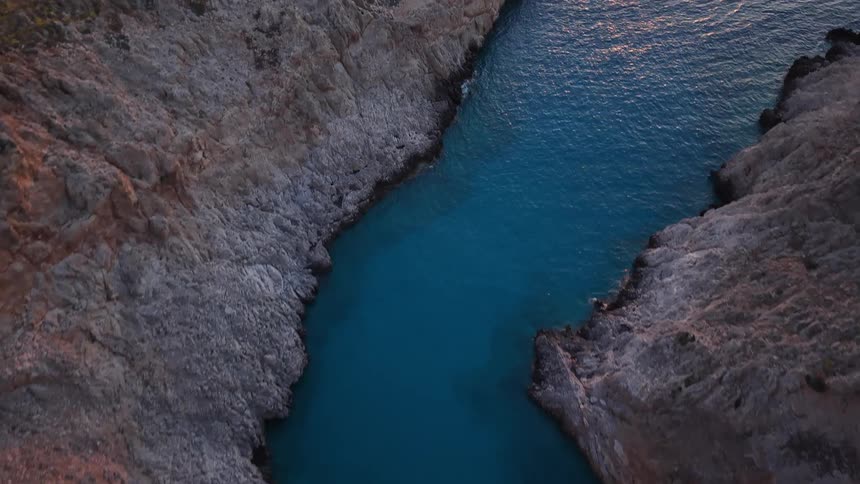 An aerial drone flies over a secluded rocky cove with turquoise water at sunset and moves out towards the open sea