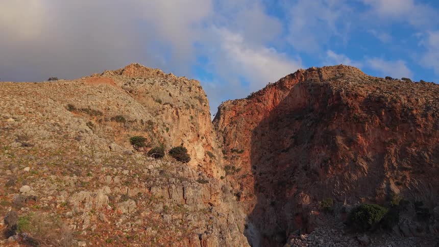 An aerial drone flies slowly towards a rugged mountain gorge as the cliffs glow with the warm light of the golden hour