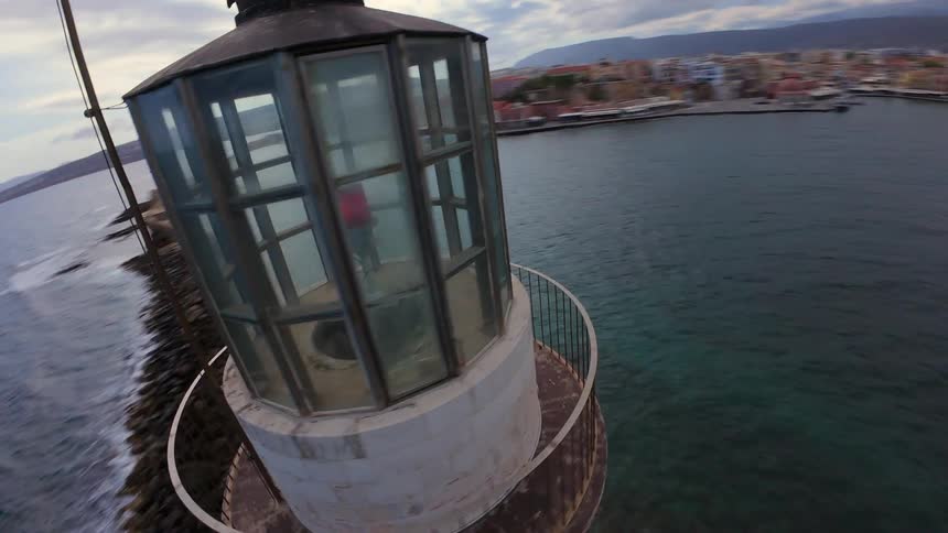 A dynamic aerial shot orbits a historic stone lighthouse as a speedboat sails past and into the harbor of a coastal town