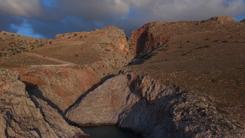 A stunning aerial view pulls back from a dramatic, rocky canyon leading down to a secluded sea inlet at golden hour