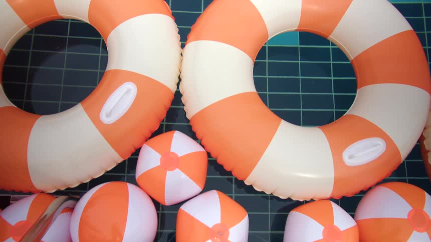 A top-down view of orange and white striped inflatable lifebuoys and beach balls floating gently in a swimming pool