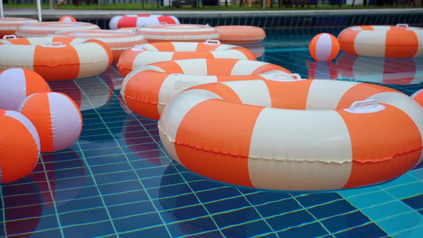 Bright orange and white striped inflatable rings and beach balls float peacefully in the still, blue water of a swimming pool