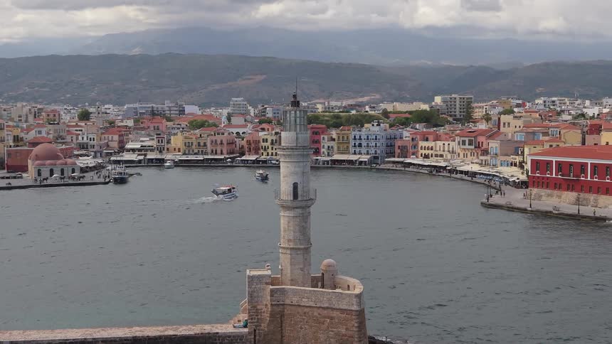 Slow-panning view of the iconic lighthouse and the old Venetian harbor in Chania, Greece, with boats in the water and a colorful cityscape along the waterfront