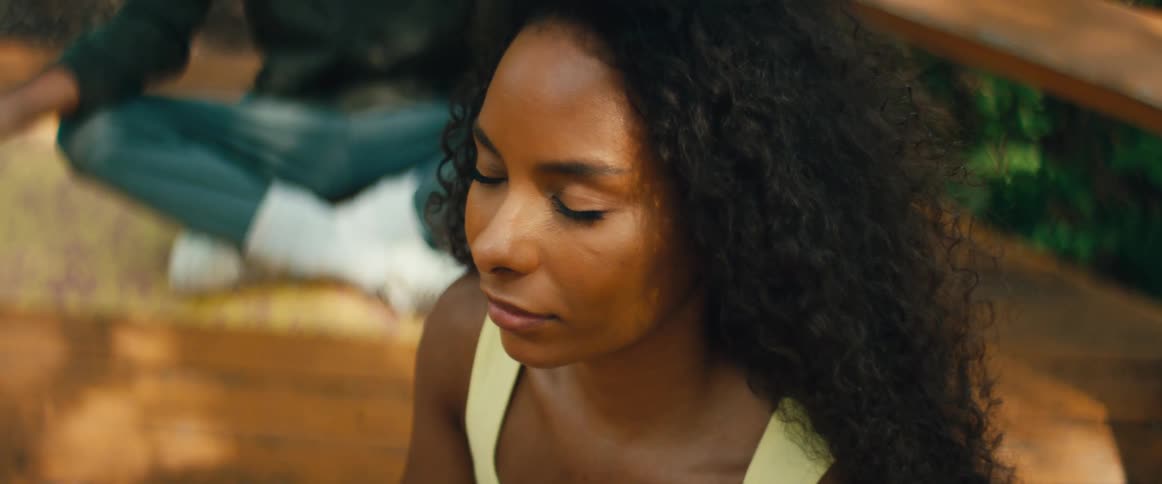 A serene woman with curly hair sits with her eyes closed in peaceful meditation outdoors