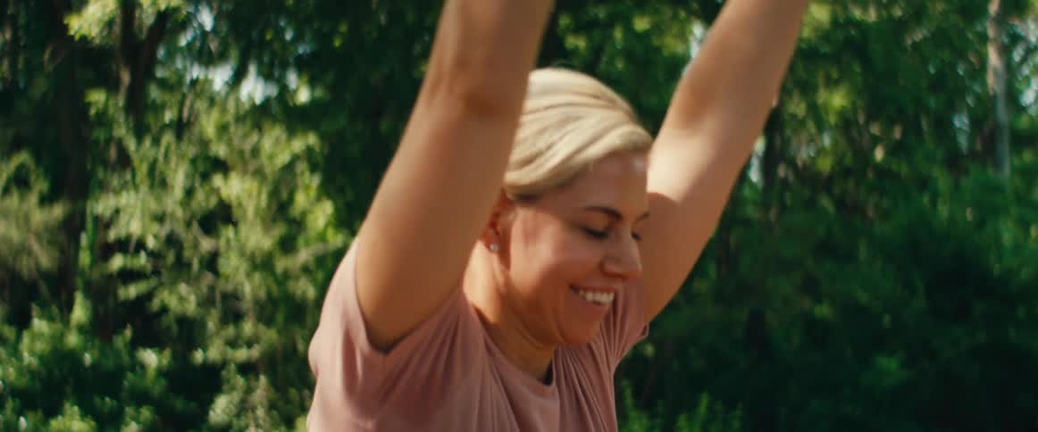 A smiling blonde woman with her hair in a bun performs a seated forward bend yoga stretch in slow motion on an outdoor wooden deck