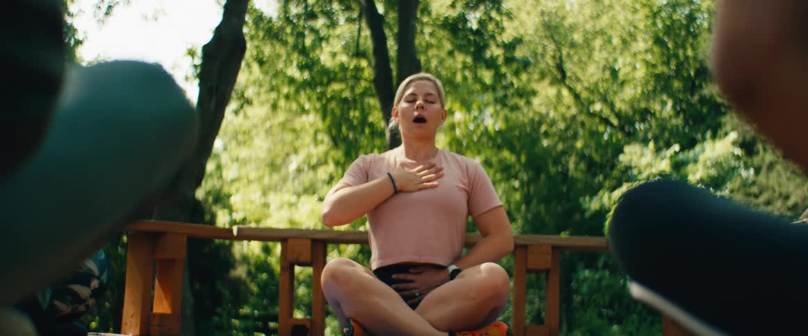 A woman practices deep breathing meditation and neck stretches while sitting on an outdoor deck surrounded by nature