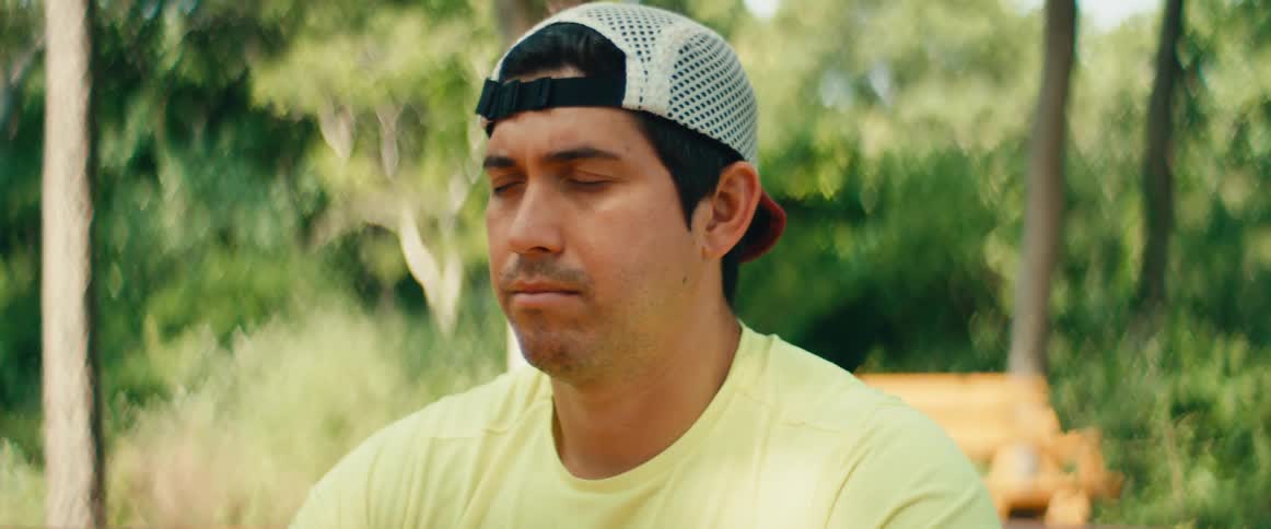 A man wearing a backwards hat and yellow shirt sits with his eyes closed, meditating peacefully in a park