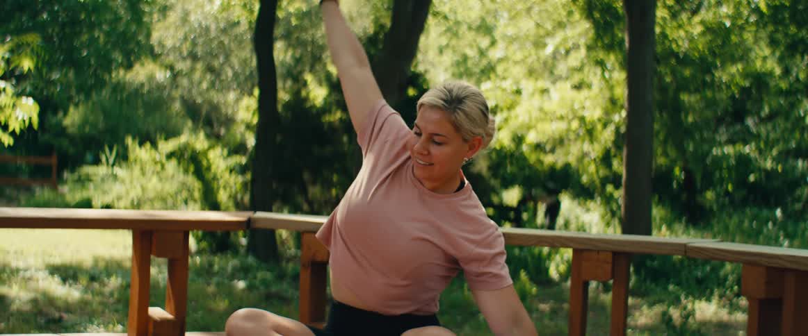 A smiling blonde woman stretches her body while doing yoga in slow motion on a sunny deck in her backyard