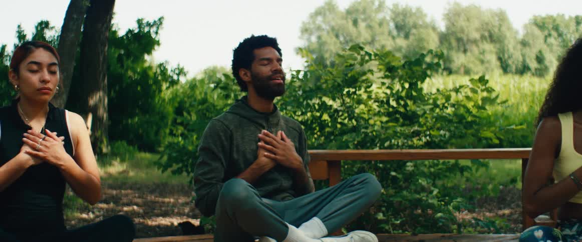 A calm and bearded man in a hoodie practices gratitude meditation with his hands on his chest while sitting outdoors in a serene, sunlit park