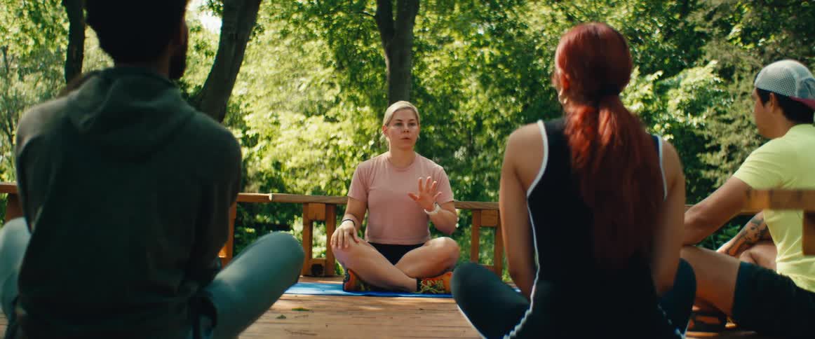 A female yoga instructor leads a group in a mindful breathing and meditation session on an outdoor wooden deck surrounded by trees