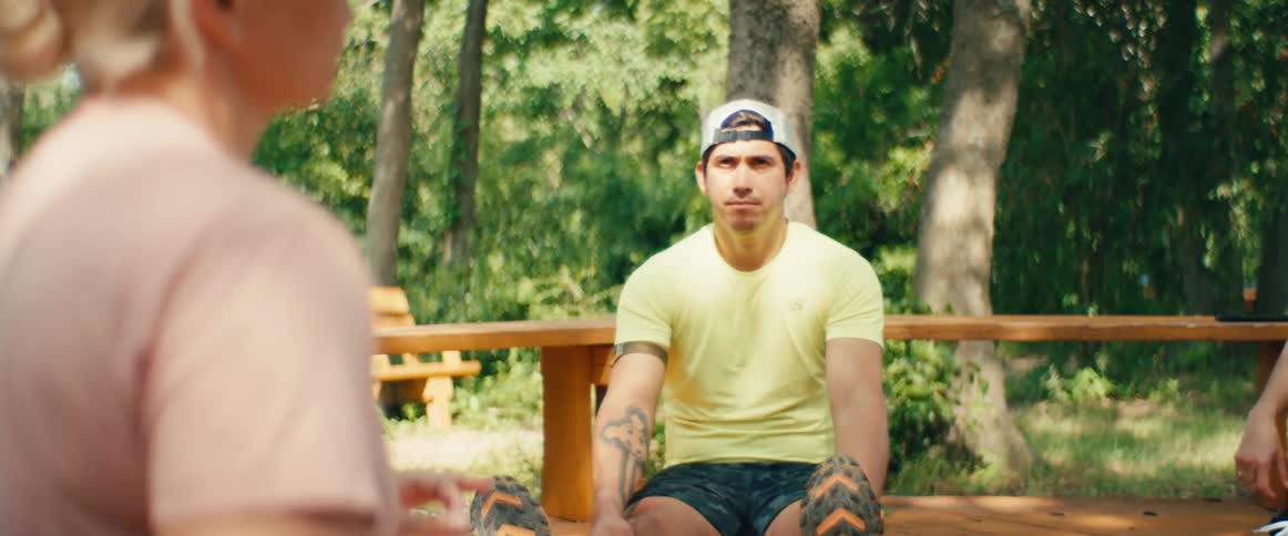 A tattooed man in athletic wear sits on a wooden deck in a park and listens to a woman speak