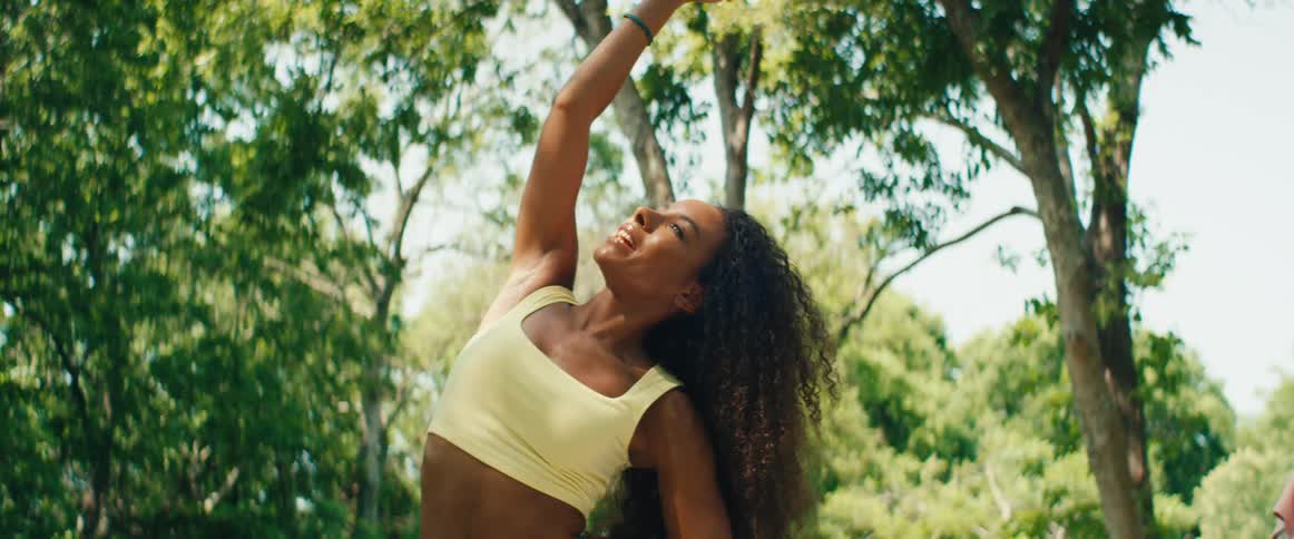 A serene woman with long curly hair performs a side stretch yoga pose outdoors in a sunlit park
