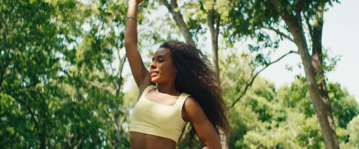 A graceful young woman with long curly hair practices yoga outdoors under the shade of lush green trees on a sunny day