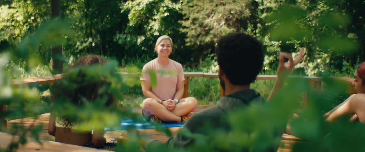 An instructor smiles as she concludes a peaceful yoga class with two students on a wooden deck in the woods
