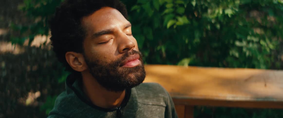 A serene man with a beard relaxes with his eyes closed, breathing deeply and enjoying the sunlight in a tranquil garden
