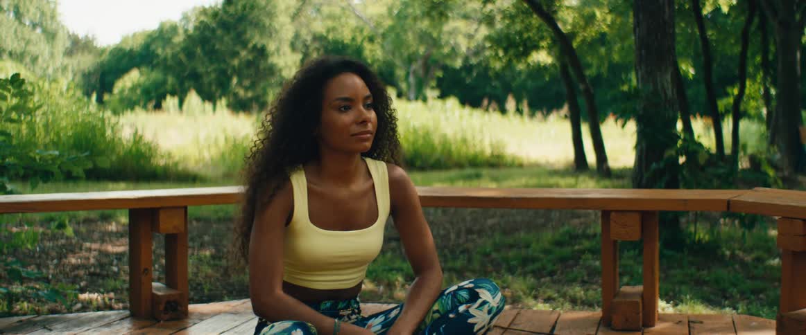 A serene woman with curly hair practices mindfulness and meditation while sitting on a wooden deck surrounded by lush greenery
