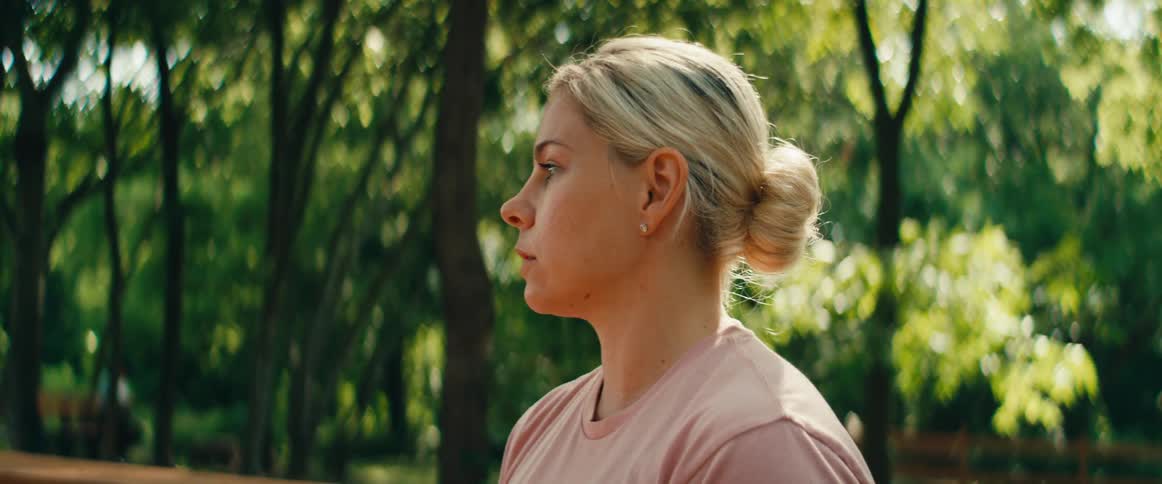 A focused blonde woman performs breathing exercises and hand gestures while meditating outdoors in a sunny park