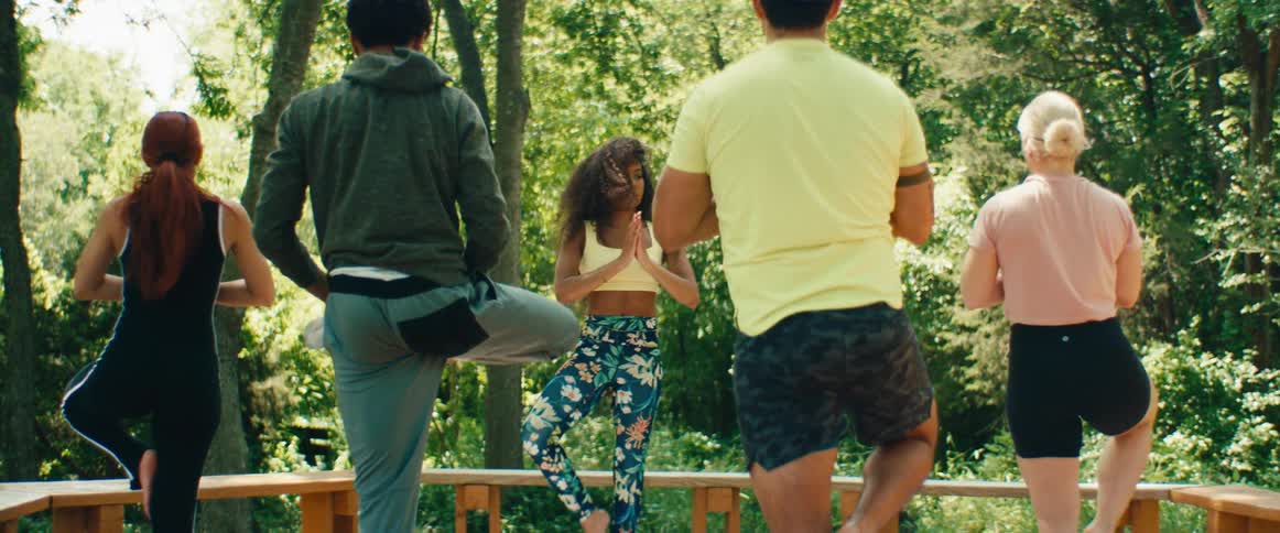A female instructor leads a diverse group of people in a balancing tree pose during an outdoor yoga class
